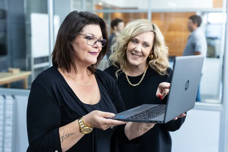 Two women smiling while looking at a laptop, one holding it and the other pointing at the screen in an office.