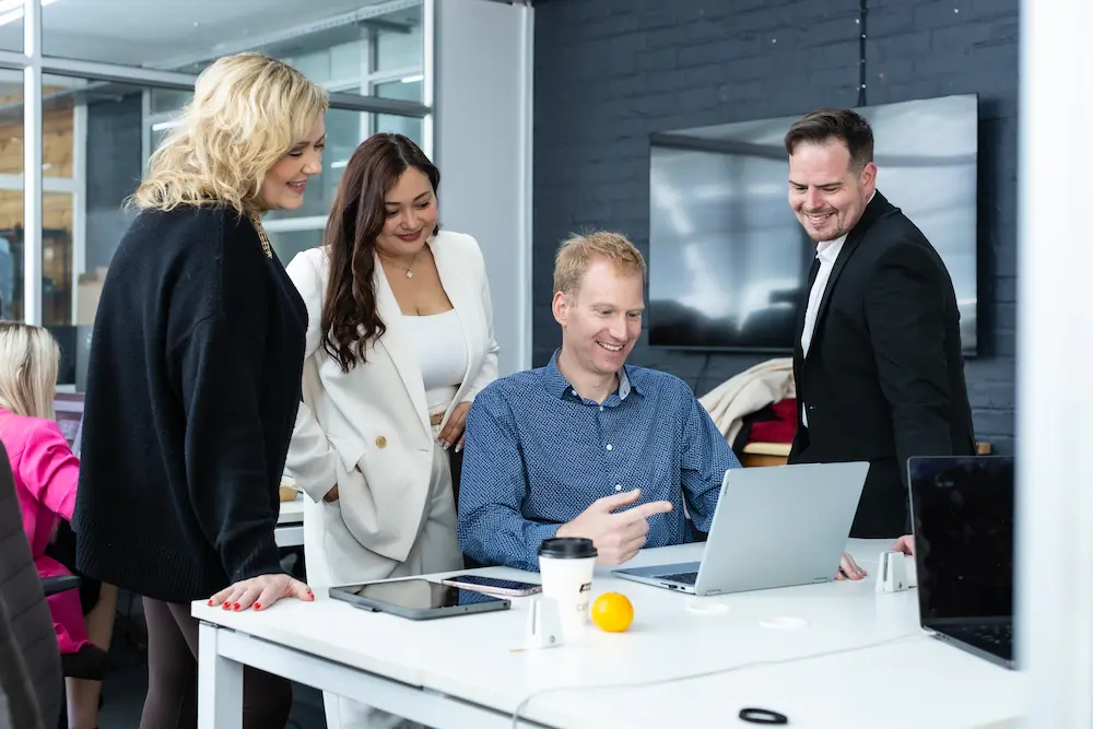Group of colleagues gathered around a desk smiling and looking at a laptop screen together in an office.