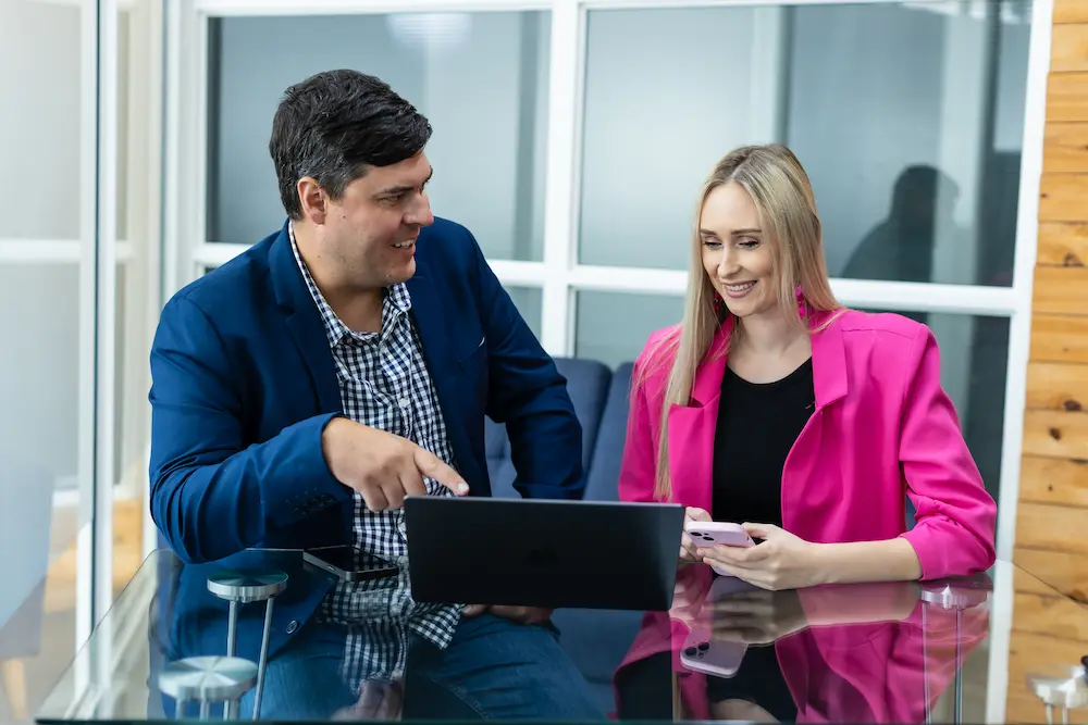 Two colleagues seated at a glass table, smiling while looking at a laptop, with one holding a phone.