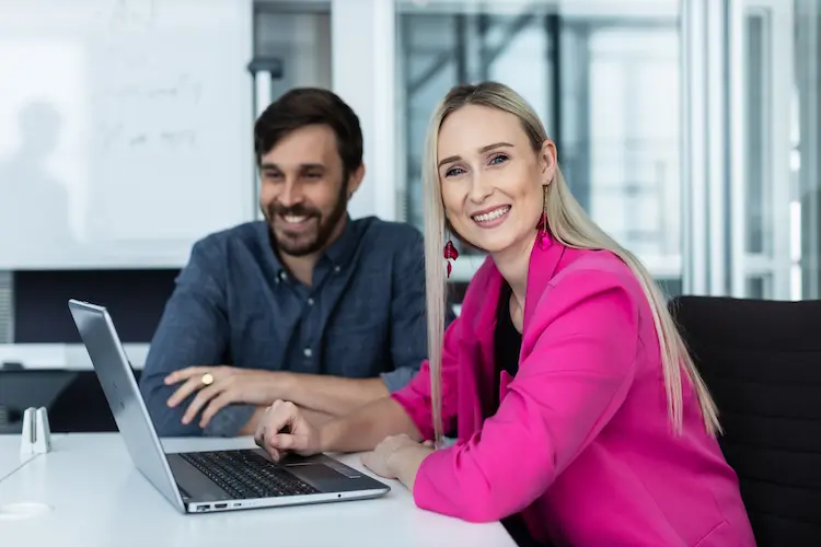Two colleagues at a desk with a laptop, woman in pink blazer smiling at camera, man in blue shirt smiling beside her.