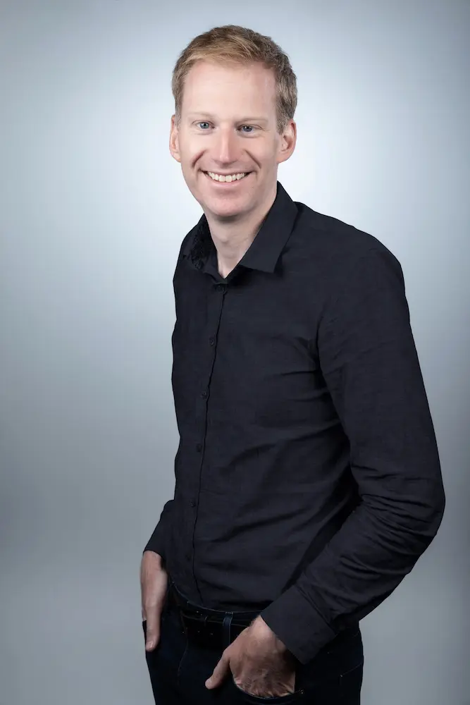 Professional headshot of a smiling person with short light hair wearing a dark shirt against a gray background.