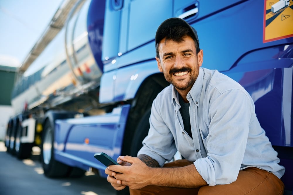 Happy truck driver texting on mobile phone while taking a break on parking lot and looking at camera. Copy space.